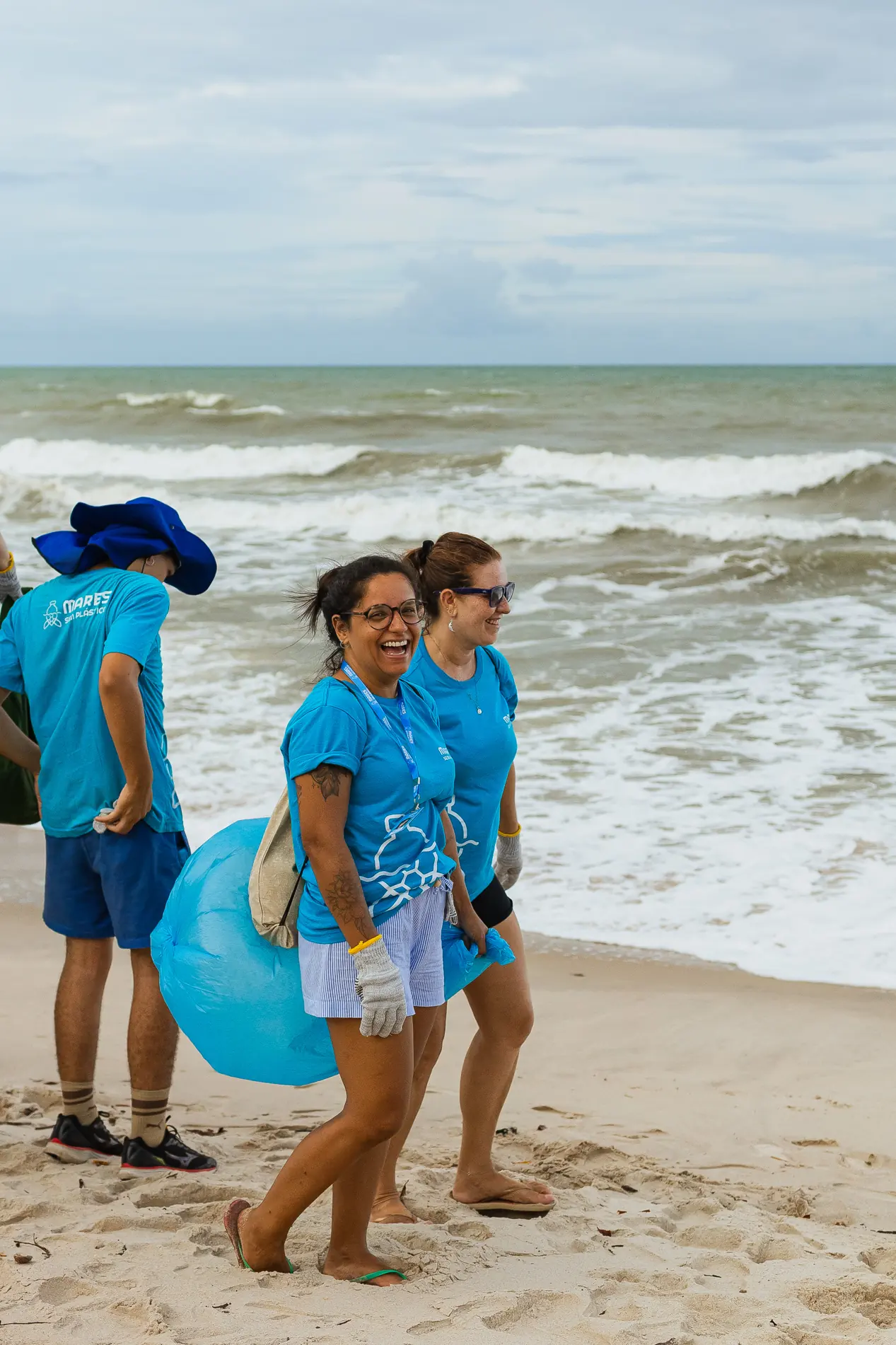 Voluntárias caminham na areia, à beira do mar, com sacos de coleta